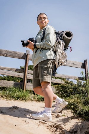 Vista de ángulo bajo de alegre joven viajera de pelo corto con mochila y equipo de viaje sosteniendo la cámara digital y mirando hacia otro lado mientras está de pie en la colina, fotógrafo de viajes