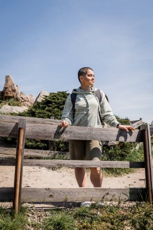 Lächelnde junge kurzhaarige Reisende mit Rucksack in lässiger Kleidung, die wegschaut, während sie in der Nähe eines Holzzaunes mit der Natur im Hintergrund steht, Wanderin beim Trekking durch die Landschaft, Sommer