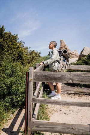 Foto de Vista lateral de la joven turista de pelo corto con mochila y equipo de viaje mirando hacia otro lado mientras está de pie en la colina con valla de madera y la naturaleza al fondo, excursionista trekking a través del paisaje - Imagen libre de derechos