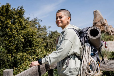 Wesoły młody krótkowłosy kobieta podróżnik z fitness tracker i plecak ze sprzętem podróżnym patrząc w kamerę, stojąc w pobliżu drewnianego ogrodzenia z naturą w tle, lato