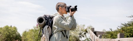 Foto de Vista lateral de la joven mujer turista tatuada y de pelo corto con mochila tomando fotos en cámara digital mientras está de pie cerca de la valla de madera en la naturaleza, caminante trekking a través del paisaje, pancarta - Imagen libre de derechos