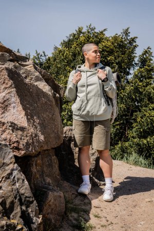 Junge kurzhaarige Touristinnen mit Fitness-Tracker und Rucksack schauen weg, während sie in der Nähe von Steinen mit Natur und blauem Himmel im Hintergrund stehen und neue Horizonte erkunden.