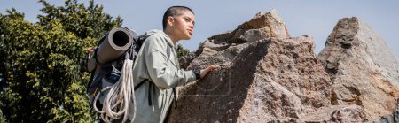 Photo for Young short haired and tattooed female hiker with backpack and travel equipment touching stone and looking away with blue sky at background, exploring new horizons, banner, summer - Royalty Free Image