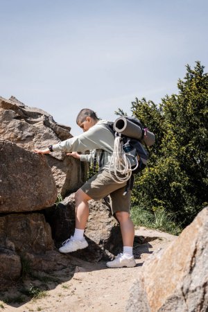 Seitenansicht einer jungen kurzhaarigen Touristin in lässiger Kleidung mit Rucksack und Reiseausrüstung, die Steine berührt und auf einem Pfad mit Landschaft im Hintergrund steht und neue Horizonte erkundet