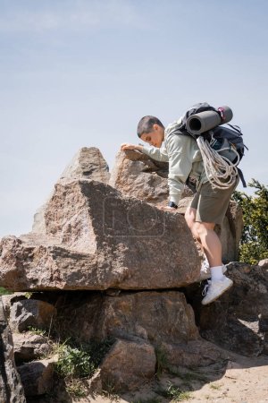 Foto de Vista lateral de una joven turista de pelo corto con mochila y equipo de viaje trepando sobre piedras con cielo azul y naturaleza al fondo, explorando nuevos horizontes, verano - Imagen libre de derechos