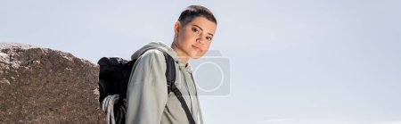 Junge kurzhaarige und tätowierte Touristin in lässiger Kleidung mit Rucksack, die in die Kamera schaut, während sie in der Nähe von Stein steht, mit blauem Himmel im Hintergrund, lebendige Reiseerlebnisse, Banner 