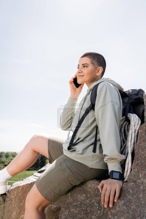 Lächelnde junge kurzhaarige Touristin mit Fitness-Tracker und Rucksack, die auf dem Smartphone pirscht und auf einem Stein sitzt, mit blauem Himmel im Hintergrund, pulsierende Reiseerlebnisse, Sommer
