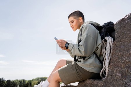 Young short haired female traveler with backpack and travel equipment using smartphone while sitting on stone with nature and blue sky at background, vibrant travel experiences, summer