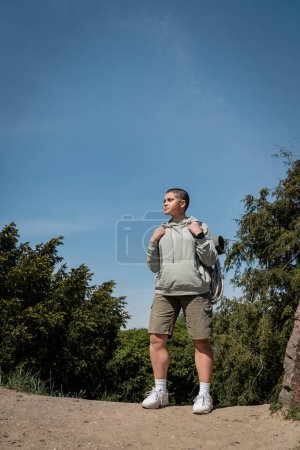 Junge, selbstbewusste, kurzhaarige Wanderin in lässiger Kleidung mit Rucksack und wegschauendem Blick im Hintergrund mit Bäumen und blauem Himmel, Übersetzung von Tätowierung: Liebe
