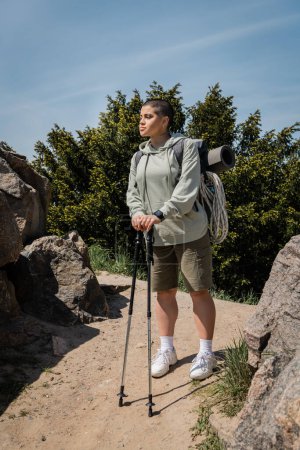 Young short haired and tattooed female hiker with fitness tracker and backpack with travel equipment holding trekking poles while standing on path with stones, trekking through rugged terrain