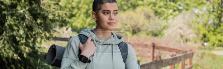 Young short haired and tattooed woman hiker with fitness tracker holding backpack and looking away and standing with landscape at background, trekking through rugged terrain, banner, summer