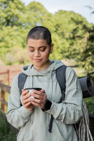 Portrait of young short haired female traveler with backpack and travel equipment holding cup of thermos and standing with blurred landscape at background, finding serenity in nature