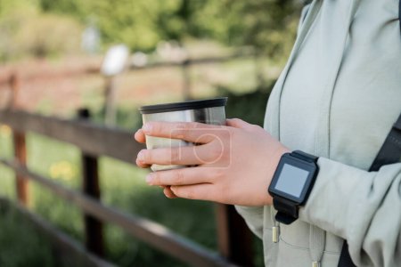 Cropped view of young woman tourist with fitness tracker holing cup of thermos and standing with blurred nature at background, finding serenity in nature, summer