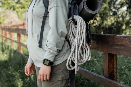 Ausgeschnittene Ansicht einer jungen Wanderin mit Fitness-Tracker, Rucksack und Reiseausrüstung mit verschwommener Landschaft im Hintergrund, die Gelassenheit in der Natur findet, Sommer
