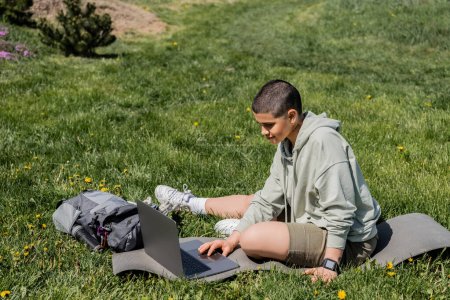 Junge kurzhaarige Touristin in lässiger Kleidung mit Laptop, während sie auf einer Fitnessmatte in der Nähe eines Rucksacks auf einer Rasenfläche mit Blumen sitzt und Gelassenheit in der Natur findet, Sommer