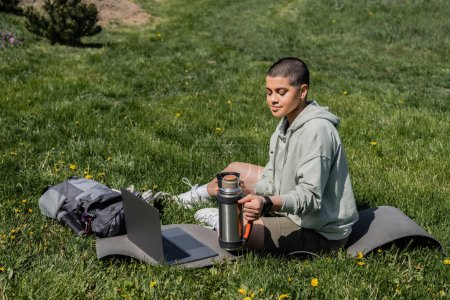Young short haired woman tourist holding thermos while sitting on fitness mat near laptop and backpack on grassy lawn with flowers, finding serenity in nature, summer, digital nomad