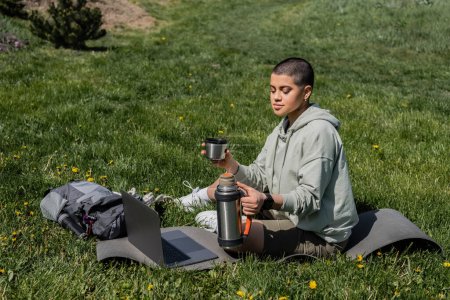Junge tätowierte und kurzhaarige Wanderin mit Thermoskanne in der Hand, während sie neben Rucksack und Laptop auf einer Fitnessmatte auf dem Rasen mit Blumen sitzt und Gelassenheit in der Natur, im Sommer, im digitalen Nomaden findet 
