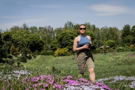 Junge kurzhaarige Touristin mit Sonnenbrille und Digitalkamera in der Nähe verschwommener Blumen mit verschwommener Landschaft im Hintergrund, Übersetzung des Tattoos: Liebe