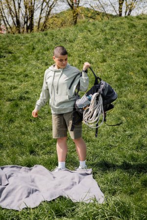 Young short haired female hiker with fitness tracker holding backpack with travel equipment while standing near blanket on grassy lawn with hill at background, connecting with nature concept