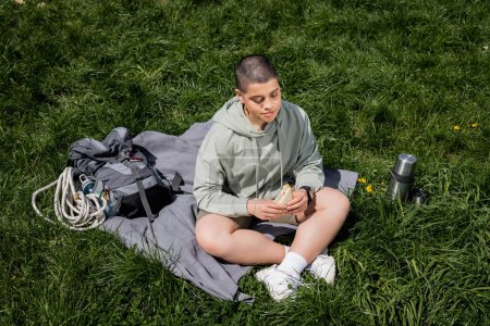 High angle view of young short haired female hiker holding sandwich while sitting near backpack and thermos on blanket and grassy lawn, connecting with nature concept, Translation of tattoo: love