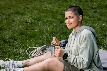 Sonriente joven de pelo corto y tatuado turista femenino sosteniendo sándwich y taza de termo mientras está sentado cerca de la mochila en la manta en el césped herboso, conectando con el concepto de la naturaleza