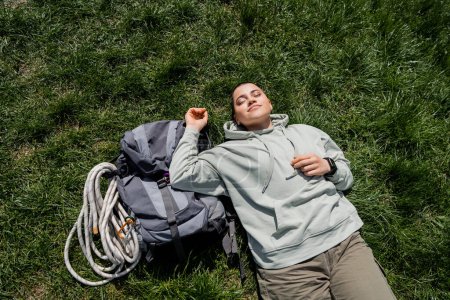 Vista superior de una joven excursionista de pelo corto con ropa casual acostada con los ojos cerrados cerca de la mochila con equipo de viaje en el césped cubierto de hierba, concepto de viaje de senderismo en solitario, verano