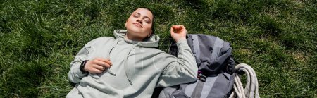 Top view of young relaxed and short haired female tourist with fitness tracker lying with closed eyes near backpack on grassy lawn, solo hiking journey concept, banner , summer