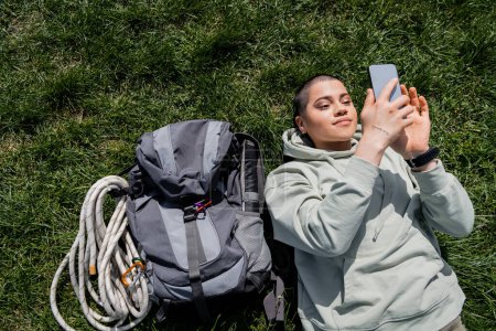 Vue du dessus de jeune voyageuse aux cheveux courts et tatouée en vêtements décontractés à l'aide d'un smartphone tout en étant couchée près d'un sac à dos sur une pelouse herbeuse, concept de voyage de randonnée en solo, été