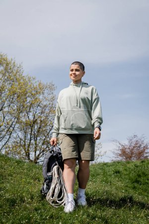 Lächelnde junge kurzhaarige Reisende mit Fitness-Tracker, der einen Rucksack mit Touristenausrüstung hält, während sie auf dem grasbewachsenen Rasen spaziert, bahnbrechend durch die malerische Landschaft, Sommer