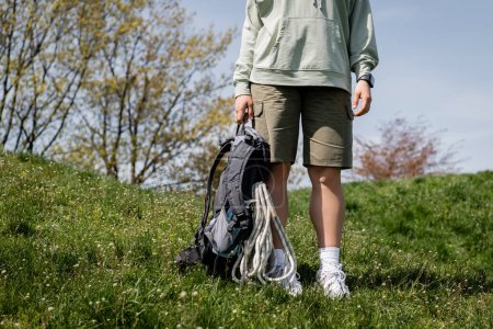 Cropped view of young female traveler in casual clothes with fitness tracker holding backpack with travel equipment standing on grassy lawn, trailblazing through scenic landscape, summer