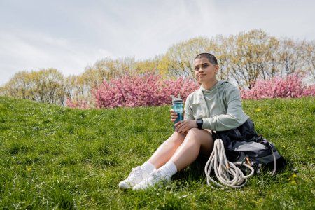 Junge, kurzhaarige und tätowierte Wanderin mit Smartwatch und Sportflasche in der Hand sitzt in der Nähe eines Rucksacks auf einem grasbewachsenen Hügel mit der Natur im Hintergrund, bahnbrechend durch die malerische Landschaft