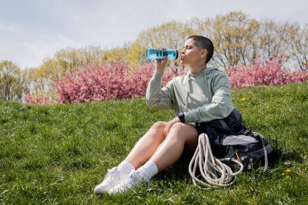 Junge kurzhaarige und tätowierte Backpackerin mit Smartwatch trinkt Wasser, während sie in der Nähe des Rucksacks auf grasbewachsenem Rasen mit der Natur im Hintergrund sitzt, bahnbrechend durch landschaftliche Landschaft