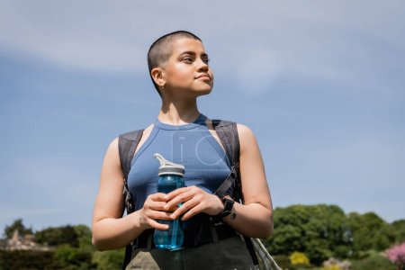 Young tattooed and short haired female hiker with backpack and smartwatch looking away and holding sports bottle with blue sky at background, trailblazing through scenic landscape, summer