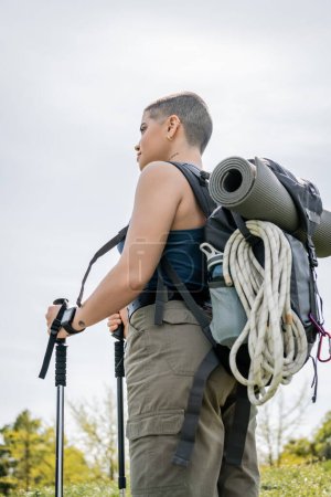 Low angle view of young short haired and tattooed female hiker with smartwatch and backpack holding trekking poles and looking away with nature at background, solo hiking journey concept