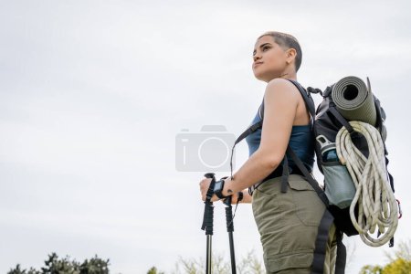 Low angle view of young short haired female backpacker with fitness tracker and backpack looking away while holding trekking poles with sky at background, solo hiking journey concept 
