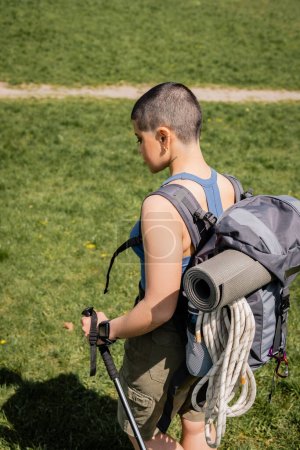 Téléchargez les photos : Vue latérale d'une jeune randonneuse aux cheveux courts et tatouée avec sac à dos et tracker de fitness tenant un bâton de trekking tout en marchant sur une pelouse herbeuse en arrière-plan, concept de voyage de randonnée en solo - en image libre de droit