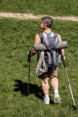 Foto de Vista lateral de una joven turista de pelo corto con mochila y equipo de viaje que sostiene bastones de trekking mientras camina en un prado herboso al fondo, concepto de viaje de senderismo en solitario, verano - Imagen libre de derechos