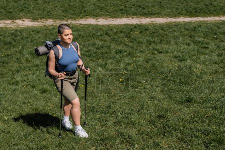 Vista de ángulo alto de sonriente joven excursionista femenina de pelo corto con mochila sosteniendo bastones de trekking y caminando sobre césped cubierto de hierba en el fondo, concepto de viaje de senderismo en solitario, verano