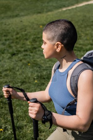 Side view of young short haired and tattooed female tourist with backpack holding trekking poles and walking on grassy lawn during summer, solo hiking journey concept