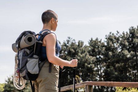 Junge kurzhaarige Touristinnen in lässiger Kleidung mit Rucksack und Reiseausrüstung, die Trekkingstöcke in der Hand halten, während sie im Hintergrund mit der Natur stehen.