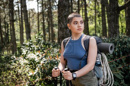 Young short haired female tourist with backpack holding trekking poles and looking away while standing in blurred green forest, reconnecting with yourself in nature concept, summer