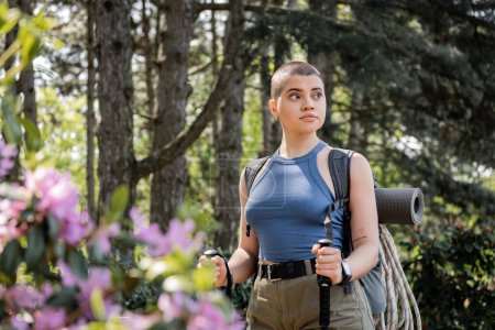 Young short haired and tattooed female hiker with backpack and travel equipment holding trekking poles while walking in green forest, reconnecting with yourself in nature concept, summer
