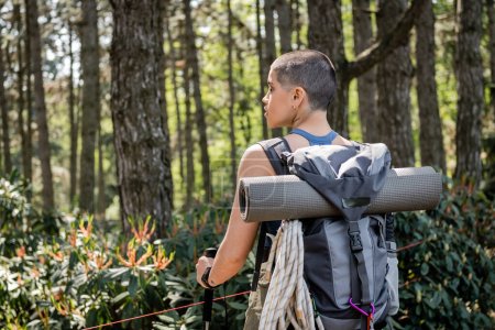 Vista lateral de la joven excursionista femenina de pelo corto y tatuada con mochila sosteniendo bastones de trekking y mirando hacia otro lado mientras está de pie en el bosque verde, reconectando con usted mismo en el concepto de la naturaleza