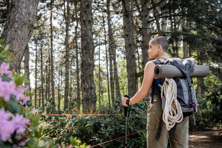 Téléchargez les photos : Vue latérale d'une jeune touriste aux cheveux courts avec sac à dos et équipement de voyage tenant un bâton de trekking et debout dans une forêt verte, renouant avec vous-même dans la nature concept, été - en image libre de droit