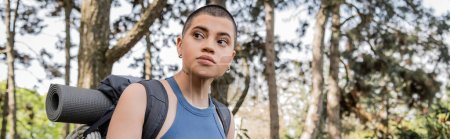 Portrait of young short haired and tattooed female tourist with backpack looking away while standing in blurred green summer forest at background, hiking for health and wellness concept, banner 