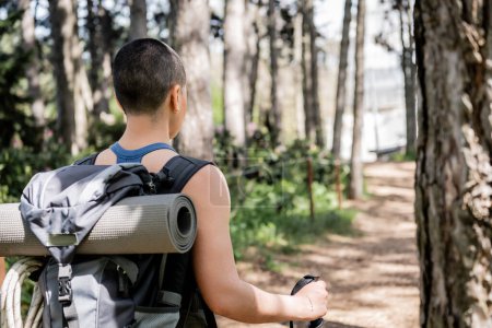Back view of young short haired and tattooed woman tourist with backpack and fitness mat holding trekking pole while hiking in blurred green forest, Translation of tattoo: love