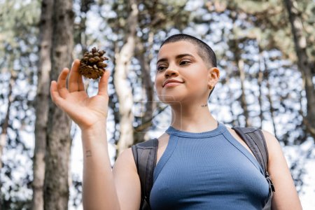 Low angle view of young tattooed and short haired female tourist with backpack holding pine cone while standing in blurred summer forest, hiking for health and wellness concept 