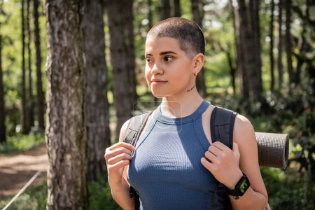Portrait of young short haired and tattooed female traveler with backpack and smartwatch looking away while standing in blurred summer forest, hiking for health and wellness concept 