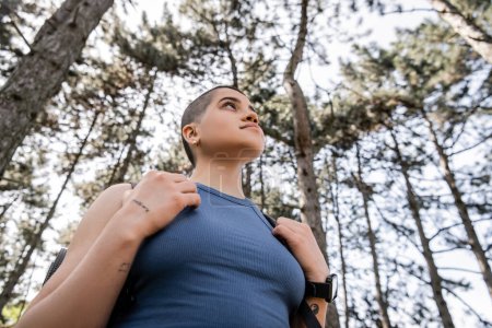 Low angle view of young short haired and tattooed female hiker with smartwatch and backpack looking away while standing in blurred summer forest, wanderlust vibes concept mug #662551490