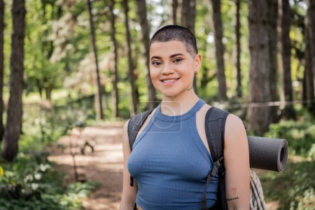 Portrait of positive young short haired female traveler with backpack looking at camera while spending time in blurred summer forest at background, wanderlust vibes concept